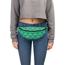 A cropped front view shows a person wearing a green fanny pack fastened around the waist. The bag features a repeating black print of a winged rod with two snakes coiled around it — a traditional medical emblem. The fanny pack is worn over blue jeans and paired with a gray cropped sweatshirt.