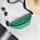 A green fanny pack with a pattern of black caduceus emblems — winged staffs with twin serpents spiraling around them — is laid flat on a pale surface. The scene includes round black sunglasses, a wide-brimmed straw hat, and a navy towel with white stripes, suggesting a relaxed or summery setting.