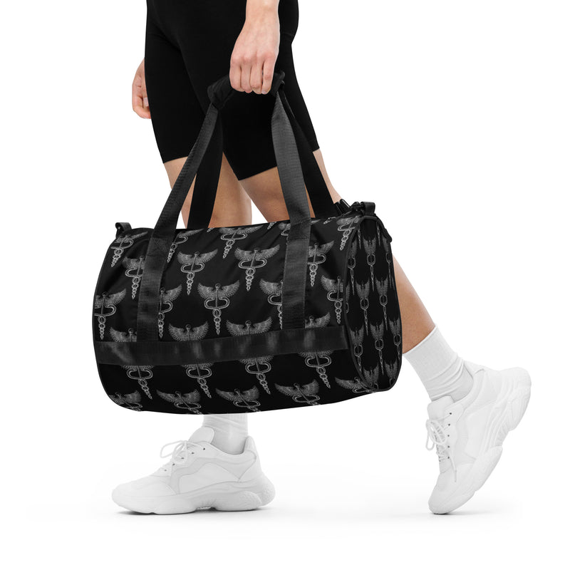 A person wearing white sneakers and black athletic wear carries a black gym bag patterned with silver caduceus symbols, each depicting a winged staff with two intertwined snakes. The bag hangs from dual handles and rests against the side of the wearer’s leg.