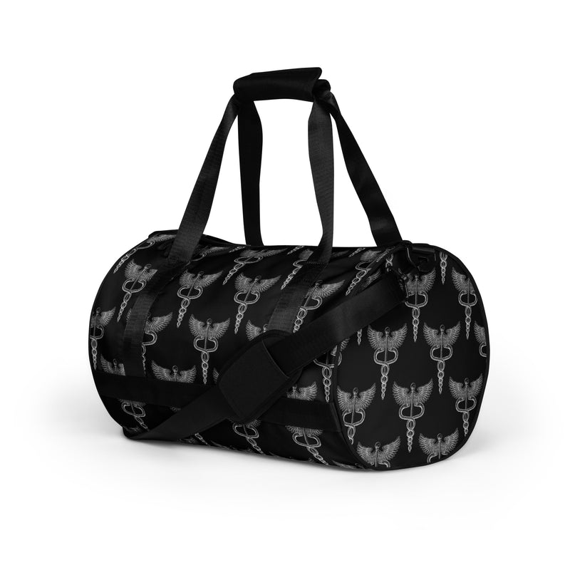 A side view of a black gym bag decorated with a repeated pattern of silver caduceus symbols—a winged staff coiled with two snakes—across the fabric. The bag has black handles and an adjustable black shoulder strap with a padded section.