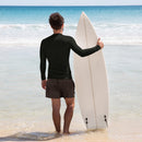 A man stands at the shoreline with a white surfboard under his arm, wearing a plain black long-sleeve rash guard and black swim trunks, facing the ocean.