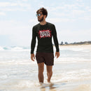 A man walks in shallow water at the beach wearing a black long-sleeve rash guard featuring a bold red graphic that reads 'BUSTED OPEN' in white, with a red splatter background. He pairs it with black swim trunks and sunglasses.