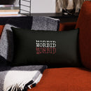 Rectangular black lumbar pillow resting on a rust-colored upholstered sofa. A black-and-white plaid throw blanket is draped behind it, and a dark shelf with stacked books and glass decor appears in the background. The pillow displays ‘MORBID’ stacked three times in white and red printed lettering, centered on the black fabric.