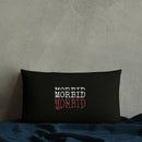 Rectangular black lumbar pillow positioned against a textured light gray concrete wall. The pillow rests on a deep blue blanket spread beneath it. Centered on the pillow, ‘MORBID’ appears stacked three times in white and red embroidered capital letters, standing out against the solid black fabric.