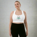 A woman standing and smiling in front of a light gray brick wall while wearing a white sports bra featuring a green and black ‘STAYING ALIVE’ design and a caduceus symbol—a winged staff with two snakes—on the front.