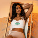 A woman reclining in a beach chair, wearing a white sports bra featuring a green and black ‘STAYING ALIVE’ logo with a caduceus symbol—a winged staff with two snakes—on the chest.