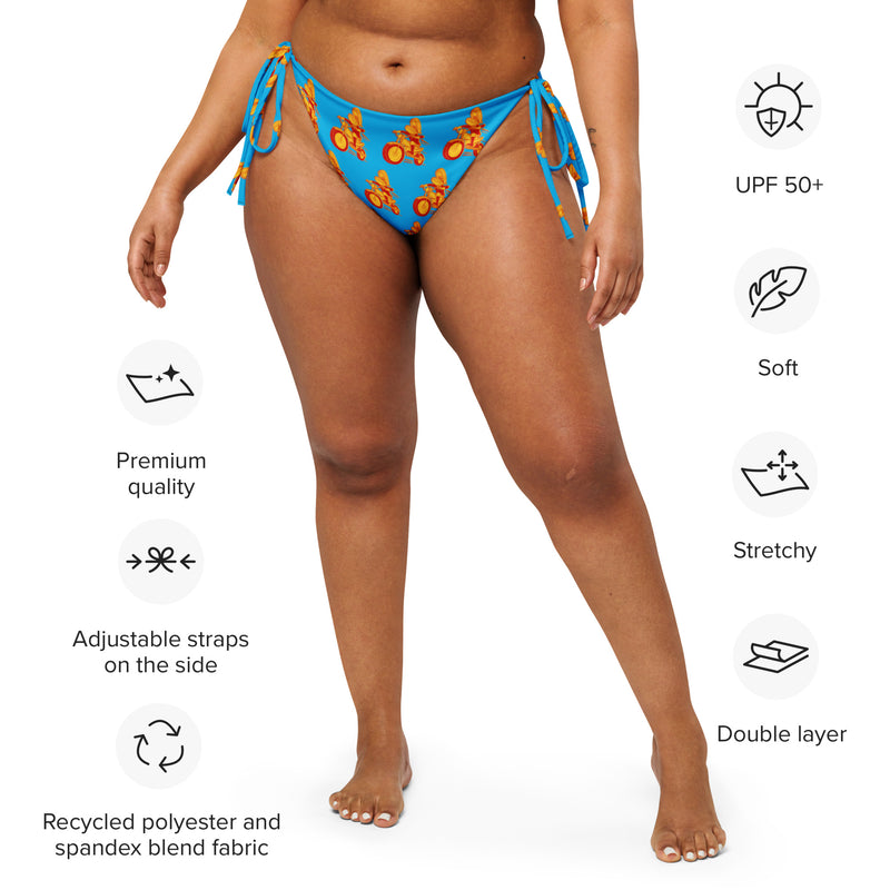 A woman models a blue string bikini bottom with a repeating motorcycle character print, surrounded by icons highlighting features such as 'UPF 50+', 'Soft', 'Stretchy', and 'Adjustable straps on the side'.
