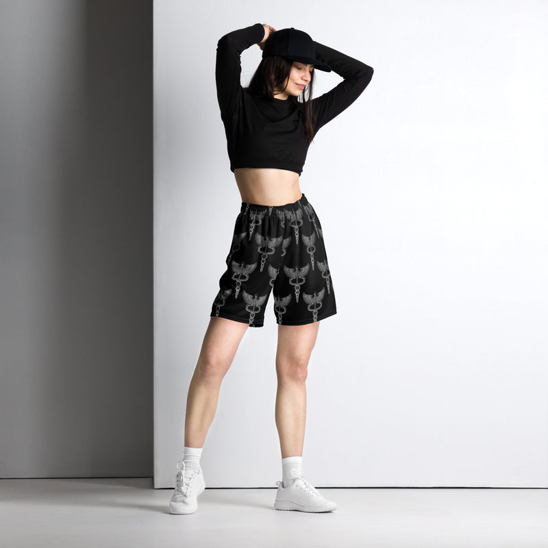 Woman poses with arms raised, wearing black mesh shorts printed with a repeating caduceus symbol—a winged staff entwined by two snakes. Paired with a black long-sleeve crop top and white sneakers.