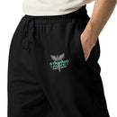 A close-up of the 'STAYING ALIVE' design on the upper left leg of the black joggers, featuring a green and gray logo and a caduceus symbol (a winged staff with two snakes) above it.