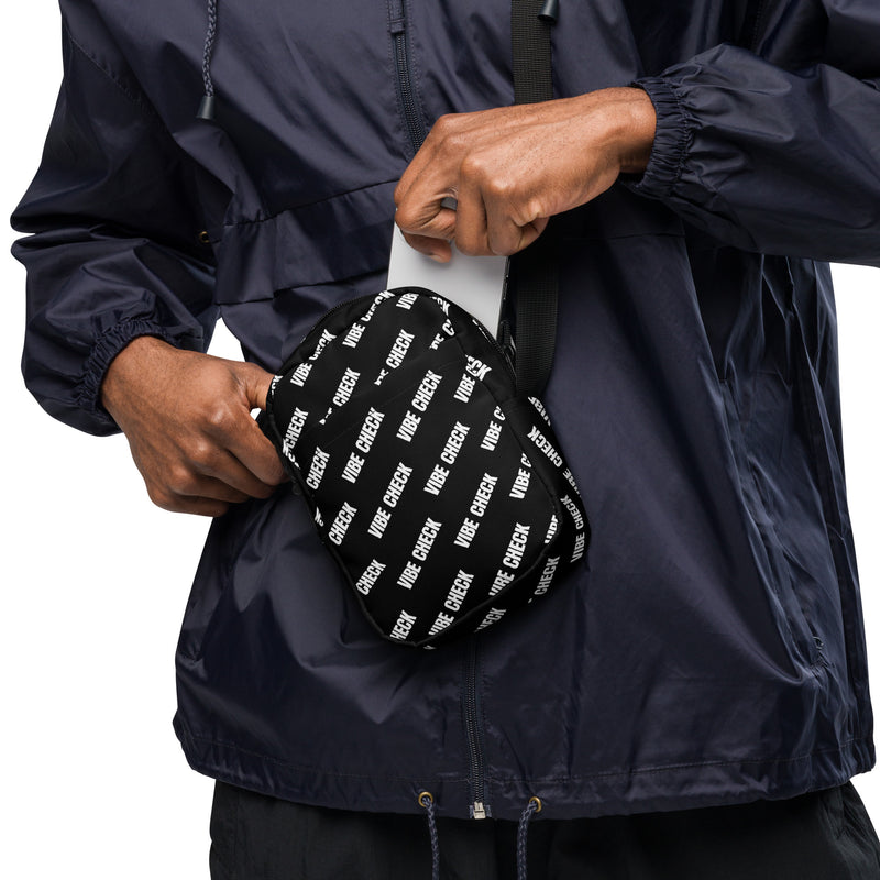 A person wearing a navy windbreaker jacket pulls a smartphone from a black crossbody bag with a white 'VIBE CHECK' print, shown slung across their chest.