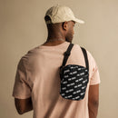 A man is seen from behind wearing a light pink t-shirt and beige baseball cap. A black crossbody bag with a diagonal white 'VIBE CHECK' pattern is slung over his shoulder and resting against his back.