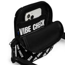 A close-up of the interior of a black crossbody bag showing a zippered compartment and inner sleeve holding a smartphone. The inner lining features the  white 'VIBE CHECK' logo across the fabric.