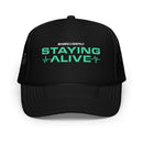 The front of a black foam trucker hat with mint green text that reads ‘STAYING ALIVE’ beneath smaller text that says ‘JON GABRUS & ADAM PALLY’, with a heart rate monitor graphic incorporated on the sides of the main text.