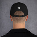 Person with short-cropped hair wearing a black dad hat viewed from behind. A white embroidered 'S' logo is centered on the back above the opening and adjustable brass buckle strap.