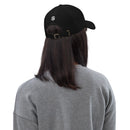 A person wearing a black dad hat viewed from the back, showing a white embroidered 'S' logo above the adjustable brass buckle strap. The person has straight, shoulder-length dark hair and is wearing a gray sweatshirt.