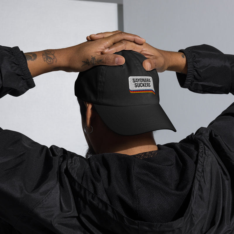 Person wearing a black dad hat with the 'SAYONARA SUCKERS' speech bubble patch on the front, sitting relaxed with hands behind head, dressed in a black windbreaker.