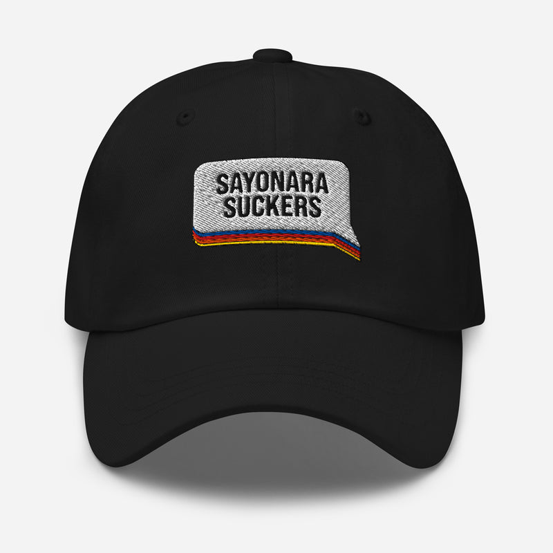 Black dad hat with a white embroidered speech bubble patch on the front. Inside the patch, bold black text reads 'SAYONARA SUCKERS' above horizontal embroidered stripes in red, orange, blue, and yellow.