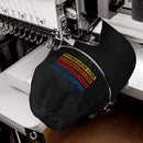 Black dad hat positioned under an embroidery machine, mid-production. The machine is stitching the 'SEXY INDIFFERENCE' design in blue, red, orange, and yellow threads.