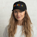 A smiling woman with long blonde hair wearing the black dad hat with the colorful 'BYEEEEEEEE' embroidered on the front, posing against a white brick wall.