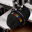 A close-up shot of a hat being embroidered on a professional embroidery machine, stitching the colorful 'BYEEEEEEEE' text on the front of the black hat.