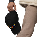 A close-up shot of a person holding the black dad hat by its back strap, showing the colorful 'BYEEEEEEEE' text on the front and the white 'S' logo on the back.