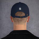 A person with a close-cropped haircut wearing a navy blue dad hat viewed from the back, showing a white embroidered 'S' logo above the brass buckle strap.