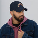 A man with a trimmed beard wearing a navy blue dad hat with a rainbow arc design of the word 'BYEEEEEEEE' on the front. He is dressed in a denim jacket over a pink hoodie, looking to the side.