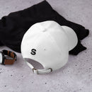 The back view of a white dad hat laid on a concrete surface, showing a black embroidered 'S' logo above the adjustable strap, next to a watch and black clothing.