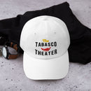 A white dad hat placed on a concrete surface next to a watch and black hoodie, with the embroidered phrase 'The TABASCO THEATER' and red pepper graphic visible on the front.