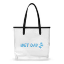 Clear structured tote bag made of transparent plastic with black faux leather shoulder straps and trim. Centered on the front is light blue text reading ‘WET DAY’ followed by a small splash graphic. The bag has an open top and flat base, allowing contents to be visible through the sides.