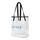 Angled front-left view of a transparent plastic tote with black shoulder straps and reinforced trim. The light blue ‘WET DAY’ text with splash icon appears on one side panel. The clear material shows seams, edges, and interior space, highlighting the bag’s structured, boxy shape.