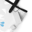 Close-up detail of the clear tote’s upper corner showing black strap attachment points, stitching, and edge trim. A thin black cord with a blank white rectangular tag hangs from the handle. The transparent plastic material and layered seams are visible, emphasizing construction and finish.