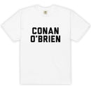 Front view of a white unisex garment-dyed heavyweight T-shirt laid flat against a light gray background. Centered across the chest in large, bold black varsity-style block letters is the stacked text ‘CONAN’ on the top line and ‘O’BRIEN’ below. The shirt has a classic crew neckline, short sleeves, and a straight hem. A small interior neck label is faintly visible at the collar.