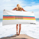 A woman stands at the shoreline holding a white beach towel featuring four horizontal stripes in blue, red, orange, and yellow, each with the phrase 'SEXY INDIFFERENCE' printed in black. The 'SiriusXM' and 'SMARTLESS' logos are printed in the bottom right corner.