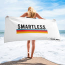 A woman stands at the shore holding a large white towel with the word 'SMARTLESS' in bold black letters above a horizontal rainbow stripe. The 'SiriusXM' and 'SMARTLESS' logos appear in the bottom right corner.