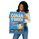 A woman wearing a light long-sleeve top and blue jeans holds a vertical blue poster featuring ‘TEAM COCO PRESENTS’ at the top and large cream text reading ‘CONAN O’BRIEN’. The script text ‘Needs a Friend’ appears below. Conan’s illustrated face is centered within a yellow sunburst near the lower right. A black banner at the bottom reads ‘FEATURING THE VOICES OF CONAN O’BRIEN × SONA MOVSESIAN × MATT GOURLEY AND YOUR FAVORITE CELEBRITY GUESTS!’.