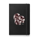 Front view of a black textured hardcover notebook with a vertical black elastic band along the right edge. Centered on the cover is a cluster of three overlapping human skulls in deep red and smoky brown tones, arranged in a triangular formation with one skull slightly forward and two behind it. The skulls show hollow eye sockets, defined nasal cavities, and detailed teeth. Beneath the cluster, the word ‘MORBID’ appears in small white capital letters on the black cover.