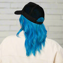 A woman with long, bright blue hair wearing a black foam trucker hat backwards, showing the mesh back and adjustable snap closure; she is dressed in a light-colored collared jacket.