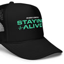 A close up, three-quarter front view of a black foam trucker hat displaying the text ‘STAYING ALIVE’ in bold mint green letters with a heart rate line below, and ‘JON GABRUS & ADAM PALLY’ in small white text above.