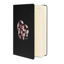 Black textured hardcover notebook shown partially open at an angle, revealing cream-colored lined pages. The front cover features three overlapping human skulls in dark red and smoky brown tones, arranged in a triangular cluster with one skull slightly forward. The skulls display hollow eye sockets and detailed teeth. The word ‘MORBID’ appears in small white capital letters beneath the design. A black elastic closure band runs vertically along the right edge.