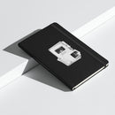 Black hardcover notebook resting at an angle across white geometric display blocks, casting a soft shadow. The textured black cover features a centered monochrome collage of layered document-style graphics. Readable text within the design includes ‘Keep it weird,’ ‘Morbid Podcast,’ ‘MORBID,’ and ‘05012018.’ A black elastic closure band runs vertically near the right edge of the notebook.
