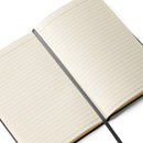 Open black hardcover notebook showing cream-colored lined pages with evenly spaced horizontal lines. A gray ribbon bookmark runs vertically down the center fold. The page edges are slightly warm-toned, and the black cover is visible along the outer margins. The interior appears clean and unused, with smooth paper and a minimalist layout designed for writing.