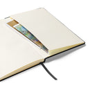 Open black hardcover notebook laid flat, showing smooth cream-colored pages and a gray ribbon bookmark extending from the bottom center. The inside back cover includes a paper storage pocket holding loose sheets and a partially visible colorful card. The pages are clean and unmarked, contrasting with the dark exterior cover and reinforcing the notebook’s minimalist interior design.