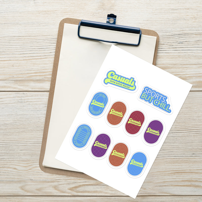 Angled top view of the 'Casuals with Katie Nolan' sticker sheet placed on a clipboard over a light wooden surface. The white rectangular sheet features two text stickers at the top—'Casuals WITH KATIE NOLAN' and 'SPORTS. BUT CHILL.'—and eight oval logo stickers in blue, red, purple, and brown tones.