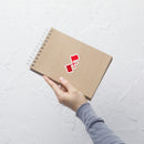 hand holding a spiral-bound notebook with a red and white logo on the cover against a textured white wall.