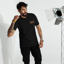 Man with a beard and tattoos wearing a black T-shirt with the phrase 'SEXY INDIFFERENCE' repeated four times in bright colors on the chest. He is standing near a silver spotlight in a well-lit studio setting.