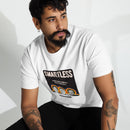 A man with tattoos and facial hair wearing a white 'SMARTLESS' T-shirt, sitting and looking off to the side in a well-lit room. The graphic on the shirt features three rainbow-outlined grayscale portraits and bold text.