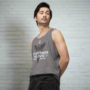 A person wearing a heather gray tank top  poses confidently in front of a white brick wall. The tank displays a caduceus graphic—a winged rod with two snakes—and bold white text that says 'STAYING ALIVE', with EKG line accents and the names 'JON GABRUS' and 'ADAM PALLY' in smaller white print.