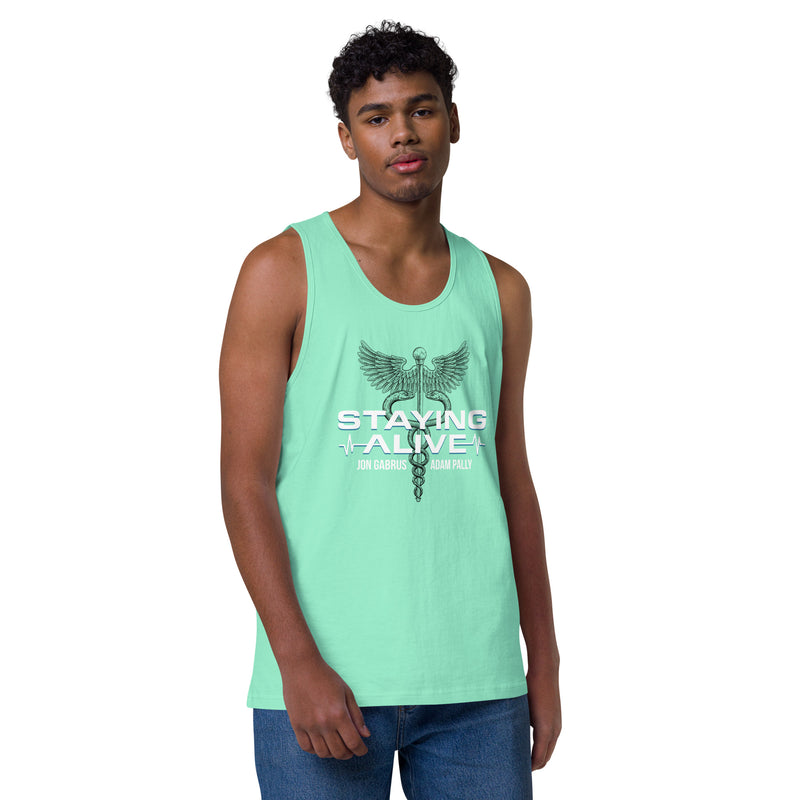A person wearing a mint green tank top and jeans stands against a plain white background. The front of the tank features a caduceus emblem—a symbol of medical care with wings and entwined snakes—above bold white text that reads 'STAYING ALIVE', with teal heartbeat lines and the names 'JON GABRUS' and 'ADAM PALLY' printed below.