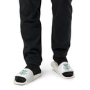 Person wearing white slide sandals with black socks. The straps display a caduceus symbol (winged staff with two snakes) above green 'STAYING ALIVE' text and the host's names in black.